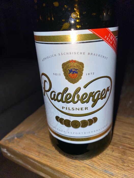 Flaska av "Radeberger Pilsner" öl, tyskt märke, etikett med kunglig sigill, grundat 1872, står på träyta.