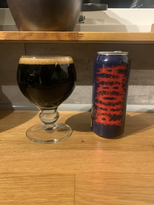 Mörkt öl i ett glas bredvid en färgglad ölburk på träbord.