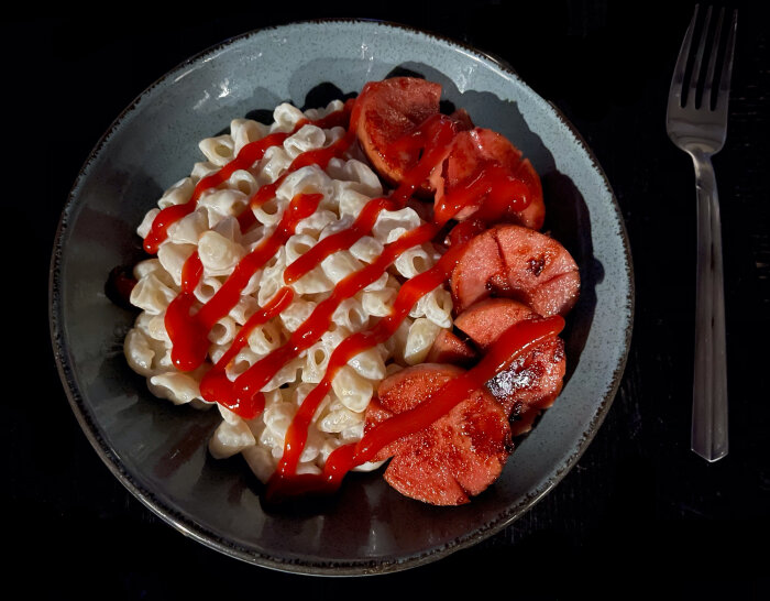 Pasta med vit sås, ketchup och grillade tomathalvor på mörk tallrik, med en gaffel.