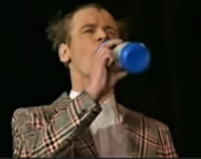 Man i rutig kostym dricker från en blå flaska på en scen. Otydlig, dålig bildkvalitet.
