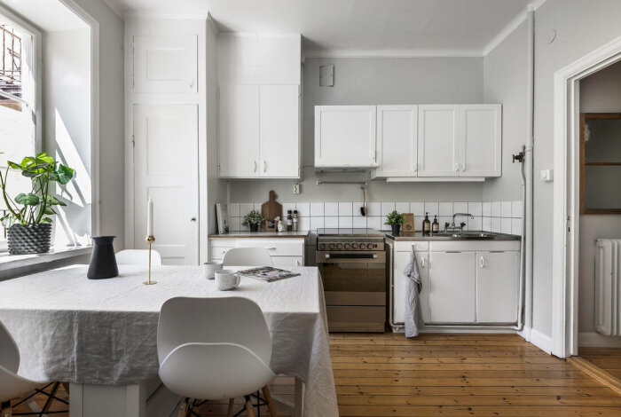 Ljust kök, vitt skåp, trägolv, modernt, bord med stolar, fönster, växter, hemtrevligt och rent.