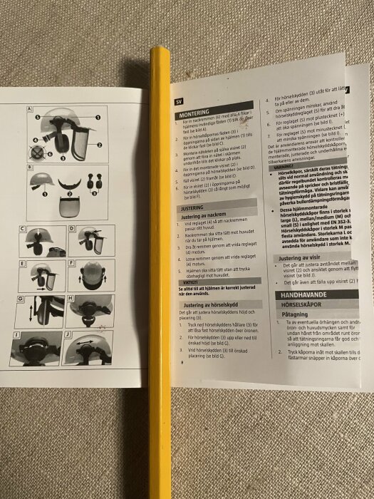 Instruktionsmanual för montering och justering av hörselskydd, öppen bok och gul penna.