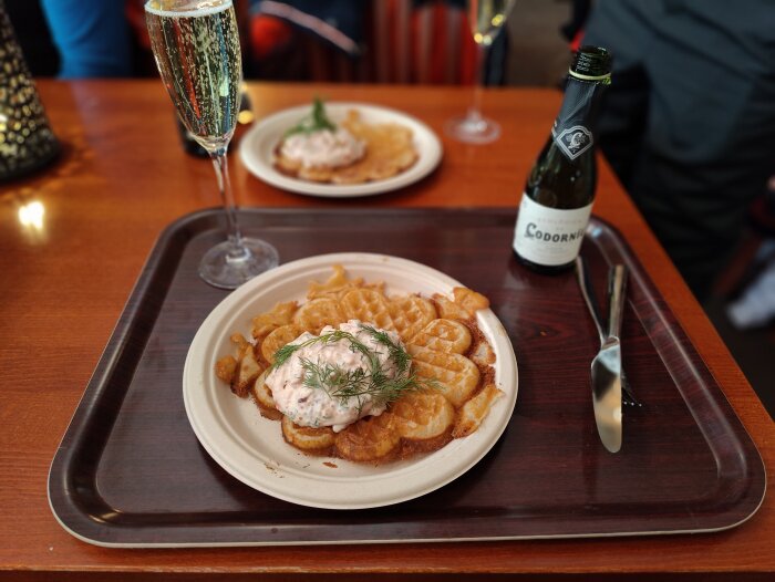 Räkmacka på tallrik, bubbel i glas, miniflaska champagne, bestick, bakgrundsaktivitet, träbord.