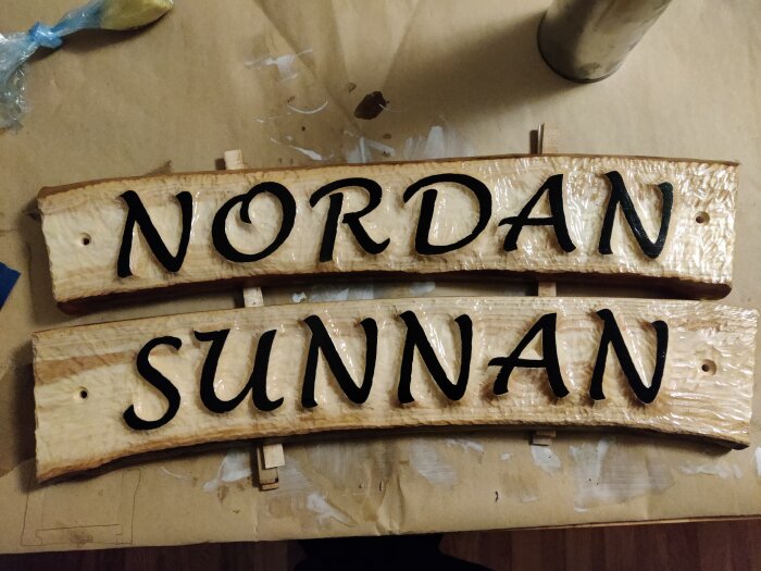 Två träskyltar med orden "NORDAN" och "SUNNAN" målade i svart. Arbetsyta med byggmaterial syns.