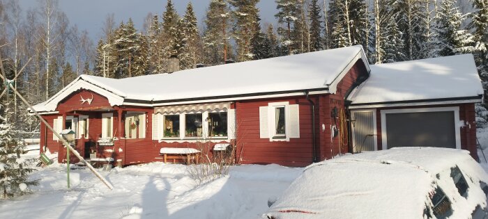Rött trähus med snötäckt tak, garage, omgivet av vinterlandskap och snöklädda träd.