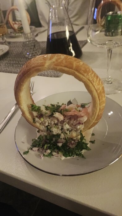 En maträtt med puffad degkrets och hackad sallad, serverat på en vit tallrik, med vin i bakgrunden.