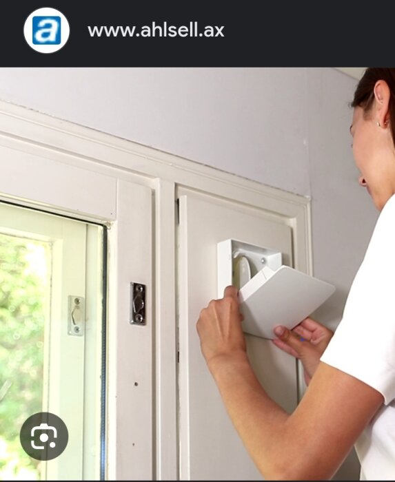 Person monterar vit, rektangulär enhet vid dörröppning; inomhus, dagsljus, teknisk installation.