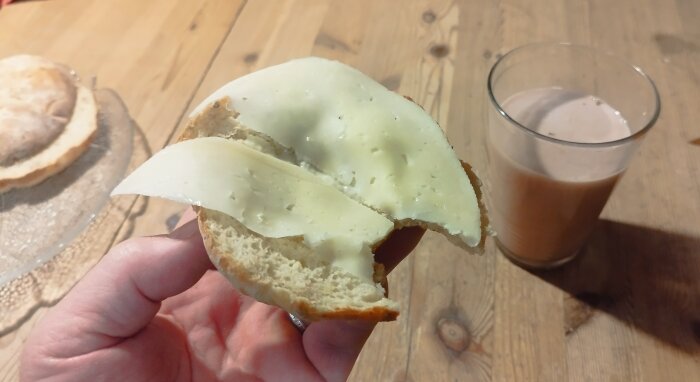 En hand håller en brödhalva med ost. Ett glas med brun dryck i bakgrunden, på ett träbord.