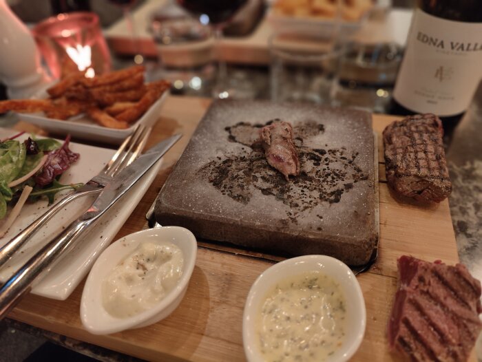 En varm stenplatta med biff, såser, sallad och sötpotatispommes på restaurangbord, rödvin i bakgrunden.