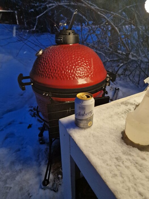 Röd grill i snö, ölburk på bord, vinterkväll, utebelysning, snötäckta träd i bakgrunden.