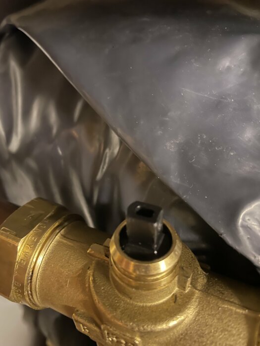 Närbild på en mässingventil och svart plastdetalj, delvis täckt av suddig grå plastfilm, mekaniska komponenter, oklar funktion.