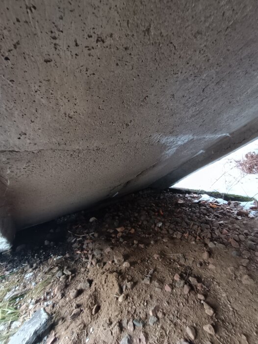 Betongkonstruktion med grus och jord under, troligtvis en bro eller kulvert, perspektivet är nära marken.