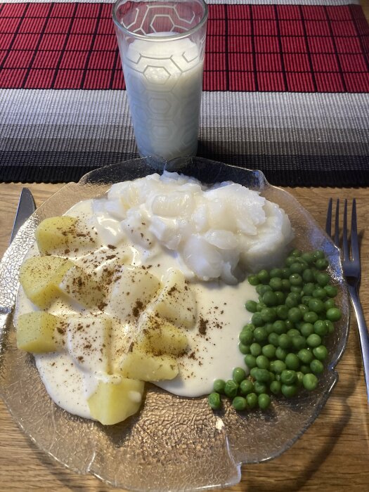 En tallrik med potatis, vit sås, gröna ärtor, och torsk. Ett glas mjölk. Röd bordstablett.