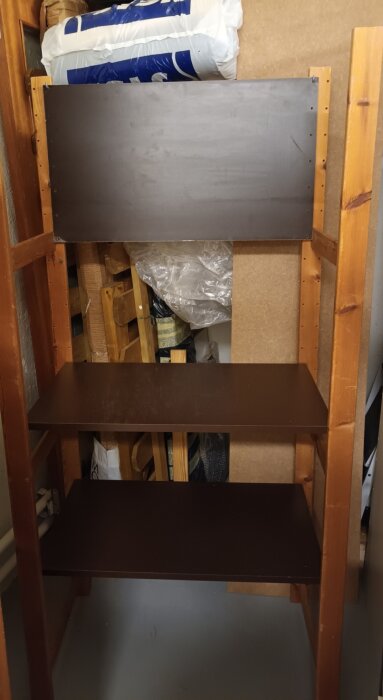 Tom trähylla i förråd med ojämnt monterad bakplatta och synliga föremål bakom.