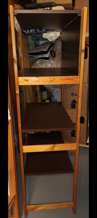 Trähylla med bruna hyllplan, står i ett rörigt förrådsrum, delvis tömd med några objekt kvar.