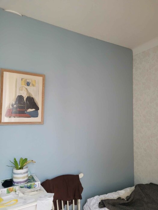Ett sovrum med blå vägg, konst, säng, kläder på stol, växt och tapet.