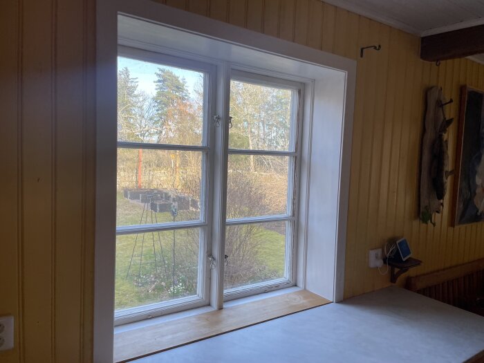 Ett rum med gula väggar, fönster med utsikt över trädgård, dagsljus, en tavla, trägolv och en bänk.