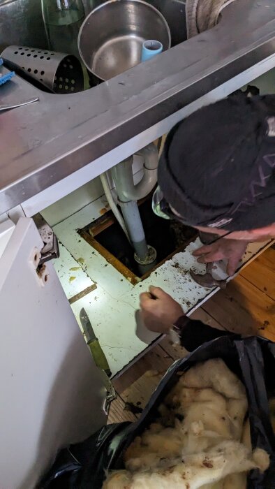 Person reparerar under diskbänk, rörledningar, öppen golvplatta, köksredskap, rörigt, renovering pågår.