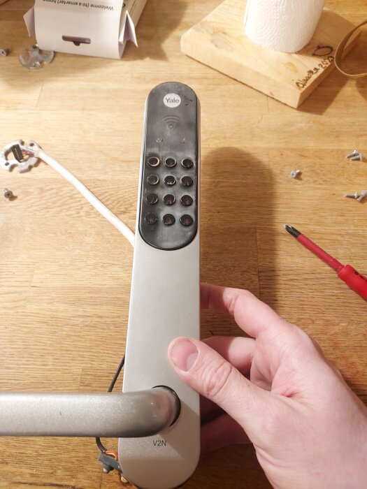 Person reparerar dörrhandtag med fjärrkontroll och verktyg på träbord.