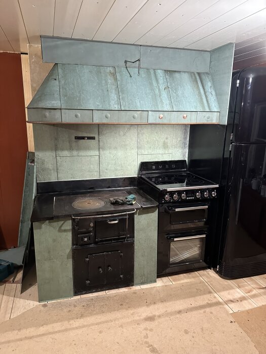Ett rustikt kök med gammal vedspis och modern svart spis, fläktkåpa, kylskåp, gröna skåplytor och vitrappat tak.