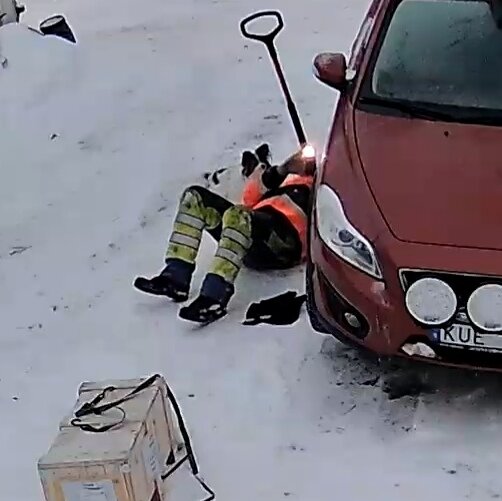 En person i arbetskläder sitter på marken vid en bil, omgiven av snö, med en snöskyffel och en låda.