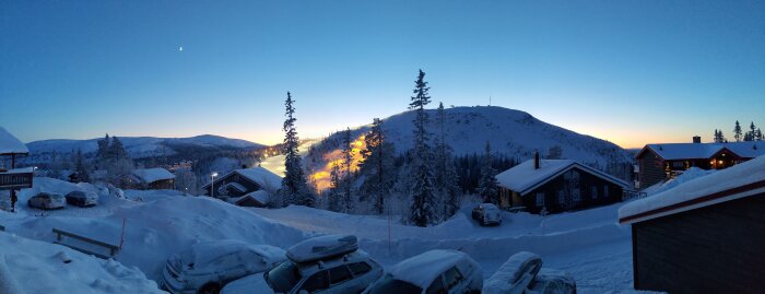 Snöiga hytter och träd vid skymning, bergslandskap, klar himmel, aftonljus, vinterskönhet, parkerade bilar under snötäcke.