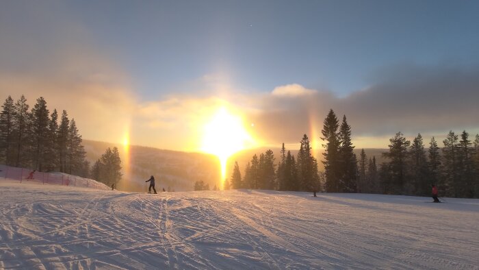 Vintersolnedgång, skidbacke, skidåkare, halo-fenomen, snötäckt landskap, träd, friluftsliv, natur, magiskt ljus.