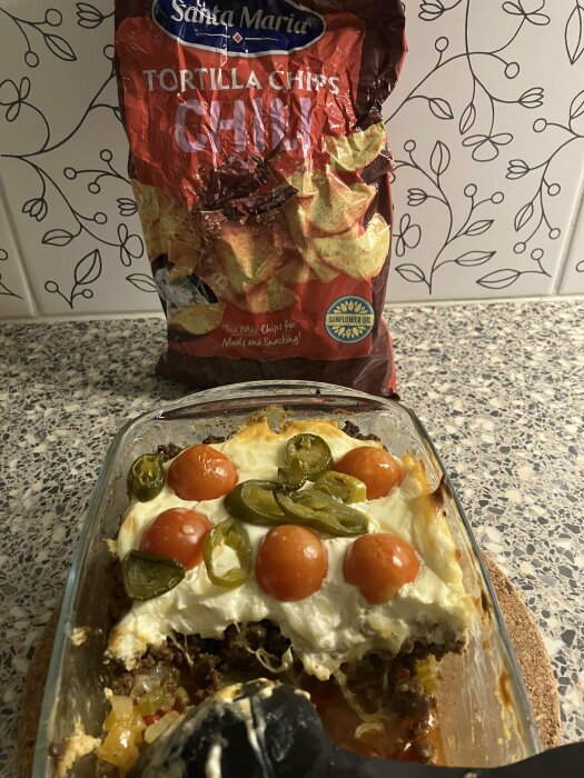 Mexikansk maträtt, troligen en form av gratäng, med gräddfil, tomater, jalapeños. Påse Tortilla Chips ovanpå, köksbakgrund.