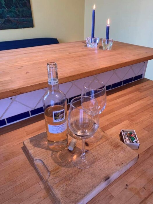 Träskärbräda med vinflaska, två glas, kork, tändstickor, tända ljus i bakgrunden.