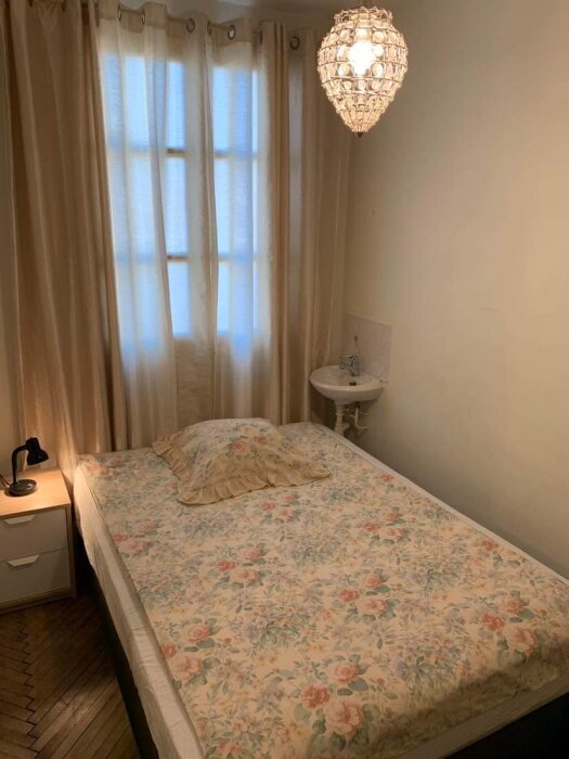 Ett enkelt sovrum med blommigt överkast, sidobord, lampa och ljuskrona. Transparenta gardiner framför fönstret.