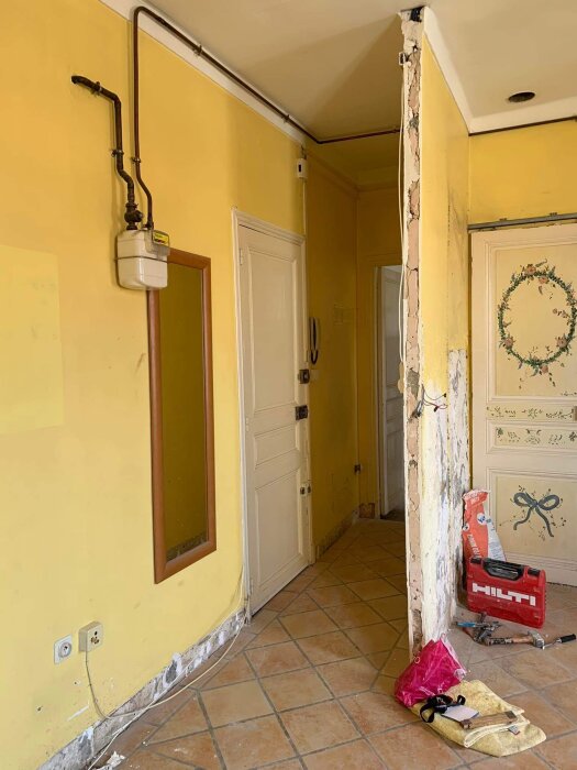 Renoveringsprojekt, skadad vägg, slitna gula väggar, verktygslåda, skor och handväska på golvet.