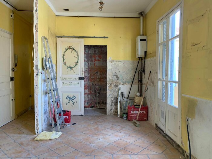 Renovering pågår i rum med skalenlig tapet, gul färg, stege, verktyg och en oavslutad vägg.