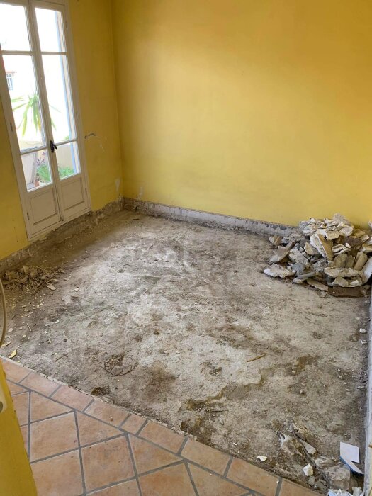 Gult rum under renovering, naket golv, rivningsavfall, tegelgolv vid dörren, vägg utan lister.