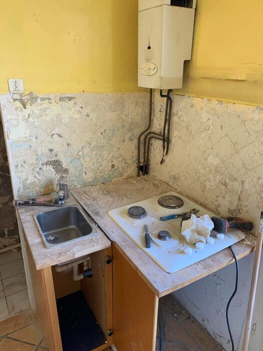 Ett slitet kök med diskho, spishäll och vattenvärmare. Behöver renovering, verktyg syns. Hygien och underhåll saknas.