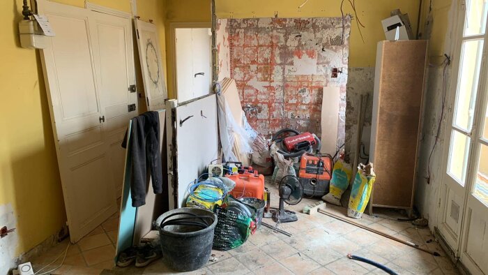 Renoveringsarbete i rum, rörigt, byggverktyg och material, avskalade väggar, golvfläkt, arbetsbelysning, ingen synlig person.