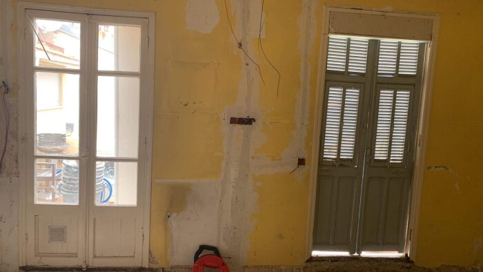 Förnstersikt från ett under-renovering-rum, gult vägg, utsatta kablar, skadad puts, persienner stängda, arbetsvakuum på golvet.