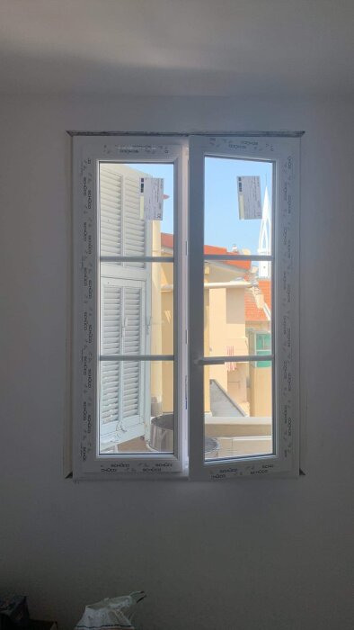 Ett fönster med öppna fönsterluckor mot en solbelyst byggnad, blå himmel syns, inomhusmiljö med skräp på golvet.