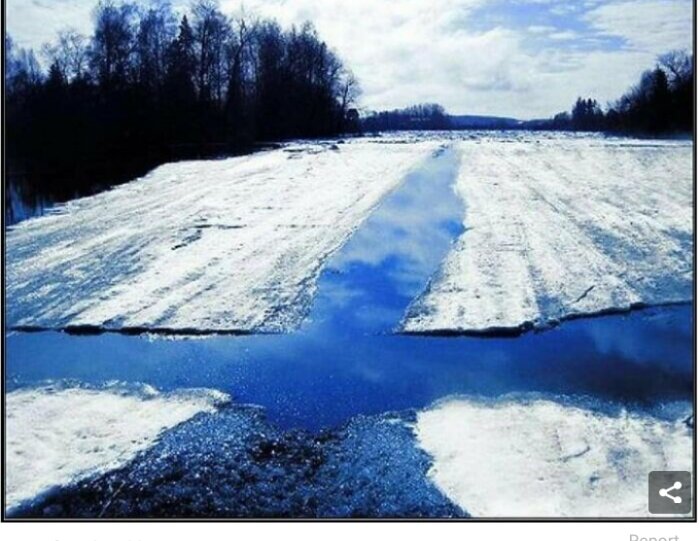 Smältande snö på isbelagd flod, blå himmel, spegling i vatten, träd vid kanter.