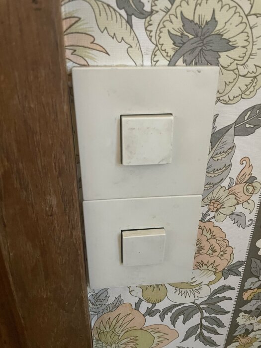 Vägg med strömbrytare, blommigt tapetmönster, synlig träkant. Slitage och smuts på brytarna.