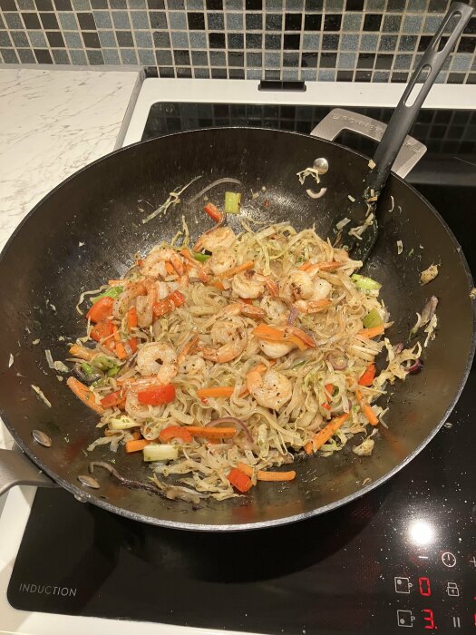 Stekpanna med räkor, grönsaker, nudlar på induktionshäll. Matlagning, stir-fry, kökeninteriör.