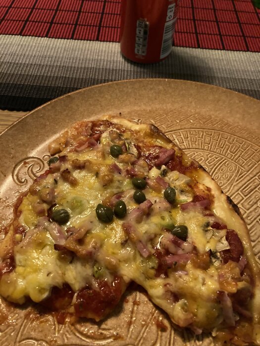 En skiva pizza med ost, skinka och gröna oliver på en tallrik, dryckesburk i bakgrunden, rödvit duk.