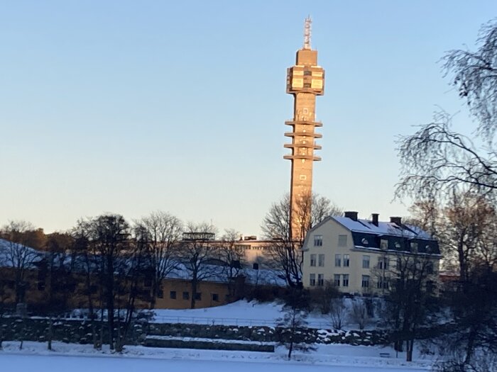 Hus och torn i snöigt landskap, blå himmel, kvälls- eller morgonsol, skuggor, vinter, ingen människa synlig.