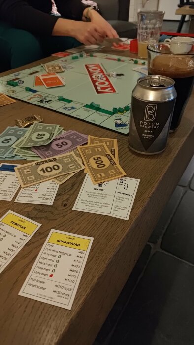 Monopoly-spelbräde med pengar, fastighetskort, tärningar, drycker och människors händer i bakgrunden.