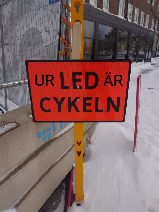 Rött skylt, "UR LED ÄR CYKELN" text, snöig omgivning, gult stolpe, byggnad i bakgrunden.