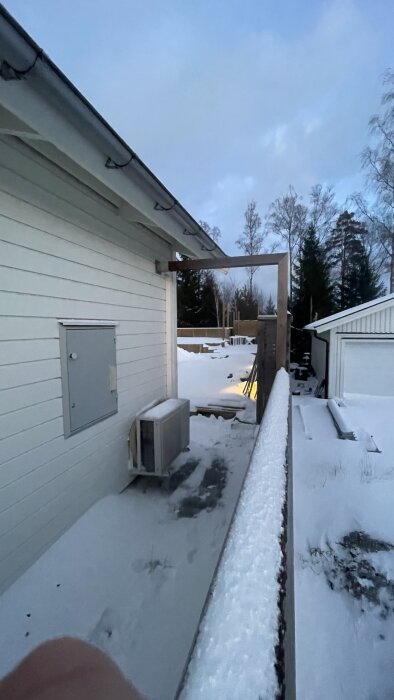 Vitklädd bakgård vid hus, snö, takränna, skymning, luftvärmepump, fotspår i snön.