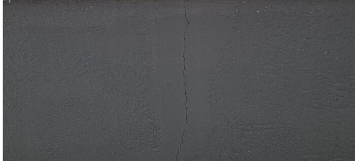 Grå betongvägg med vertikal spricka, ojämn textur, slitna detaljer, antydan till åldring och skador.