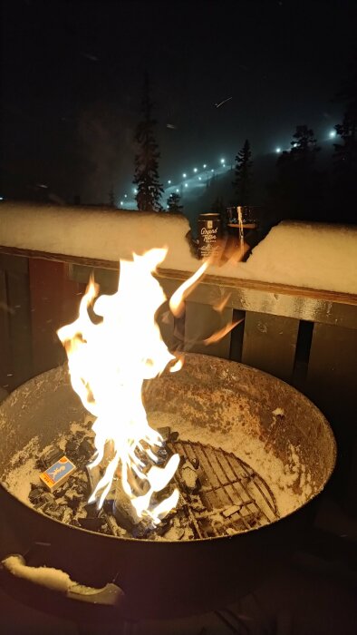 Flamma i eldkorg på en terrass med snö, natt, skidbacke med belysning i bakgrunden.