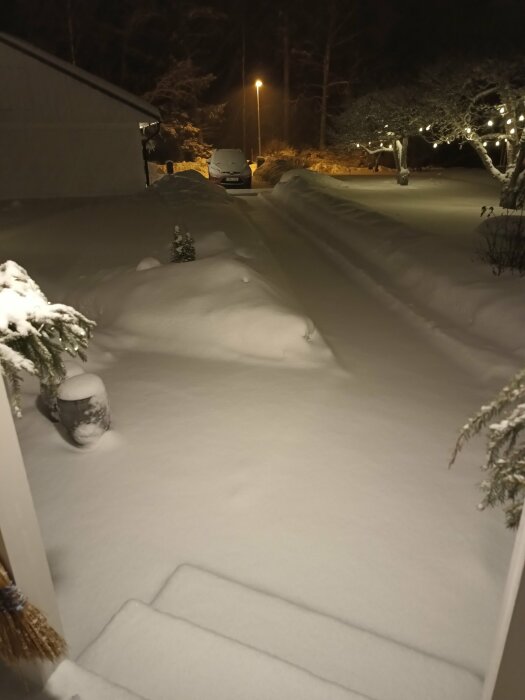 Snötäckt uppfart på natten, med lyktor, bil och träd, i tyst villaområde.