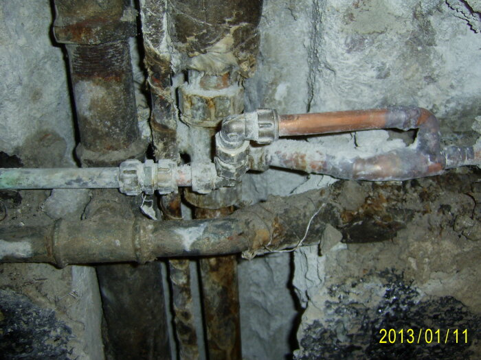 Äldre rörledningar och ventiler med korrosion, inbyggda i grov betongvägg. Kopparrör synliga. Datumstämpel i hörnet.