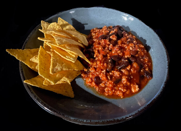 Ett fat med chili con carne och tacoskal på mörk bakgrund.